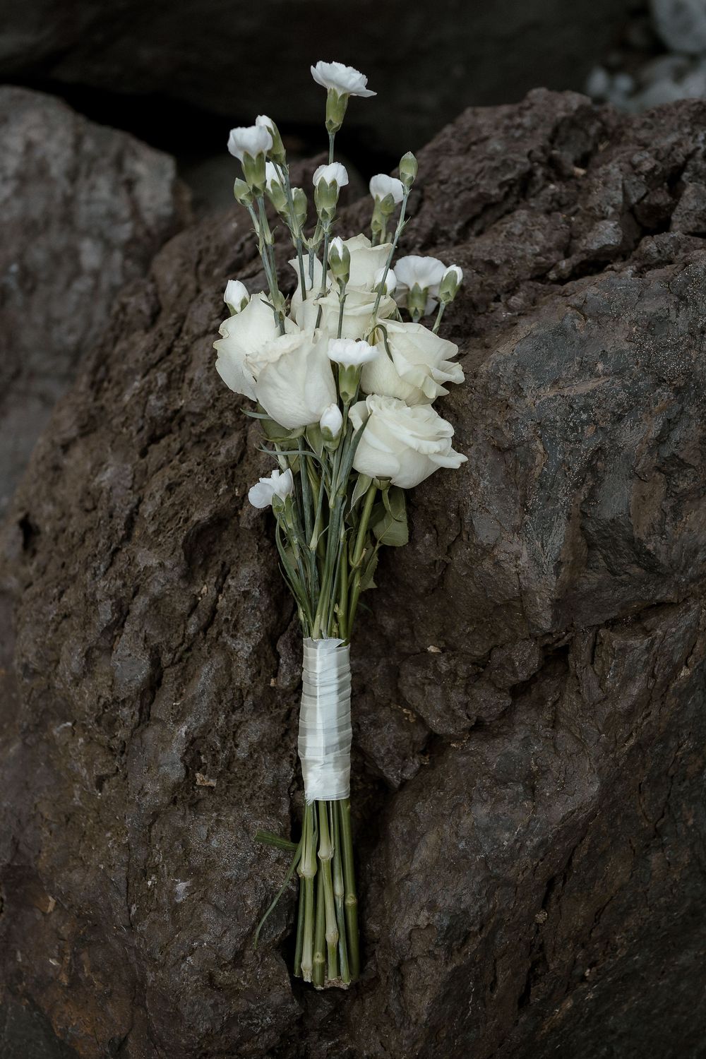 White bridal bouquet of roses and carnations wrapped in ribbon, resting against dark rocky surface.