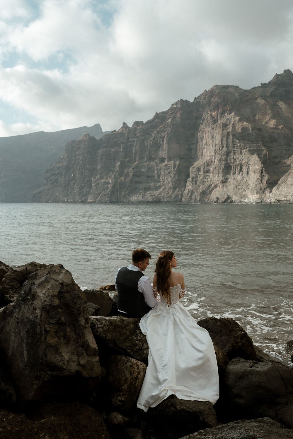 Bride and groom sitting on coastal rocks gazing at dramatic ocean cliffs on their wedding day.