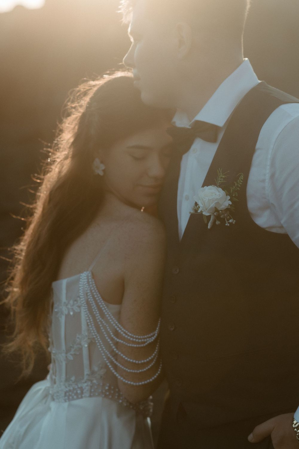 Bride and groom share a romantic golden hour moment, her pearl-draped wedding gown glowing in warm sunset light.