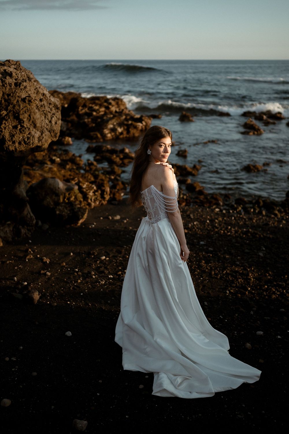 Bride in white off-shoulder gown stands on rocky black sand beach with ocean waves at dusk.
