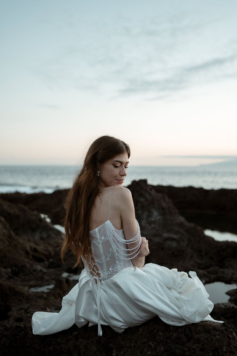 A bride in a white off-shoulder gown sits on rocky shore at dusk, her long hair flowing, ocean in background.