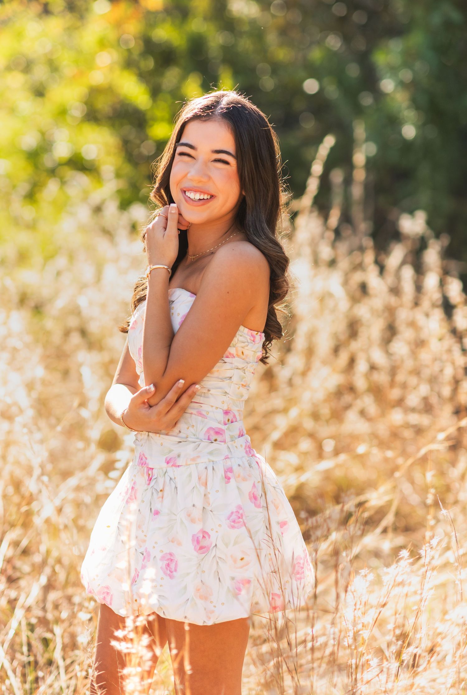 Young woman in floral strapless dress smiling outdoors in golden field with sunlit trees in background.