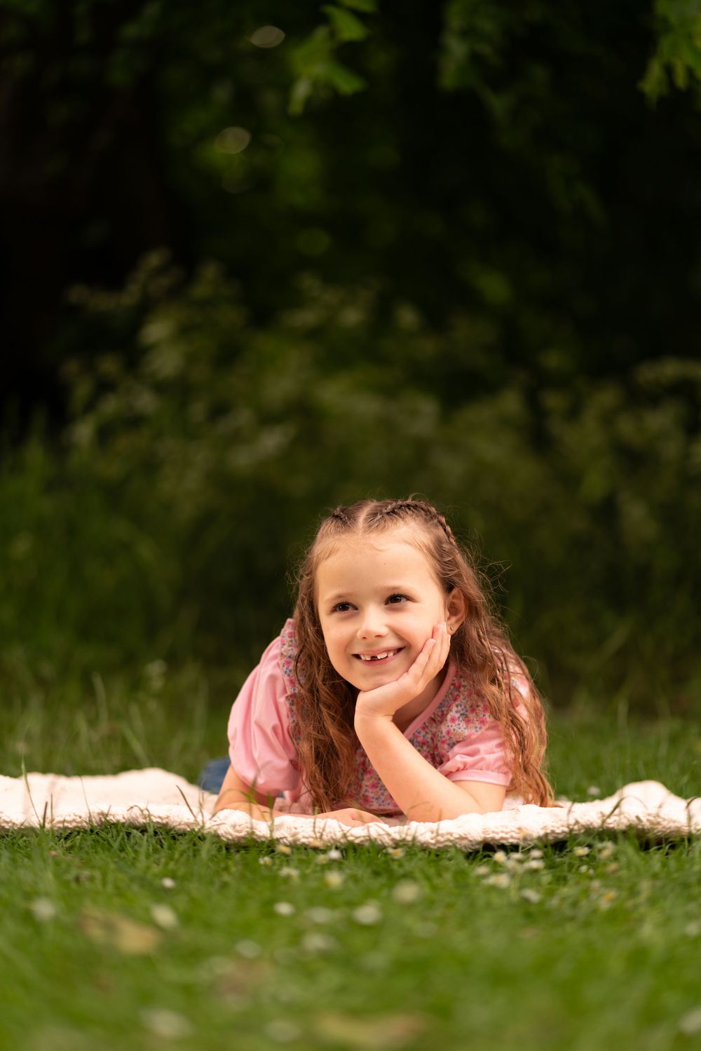 A young girl with long brown hair lying on a blanket in green grass, smiling with hand on chin during childhood photoshoot in Lincolnshire.