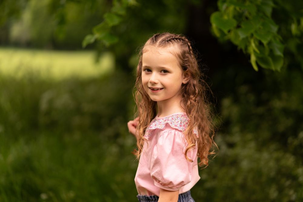 Young girl with curly hair smiles outdoors in a pink blouse, surrounded by lush green trees during childhood photoshoot in Lincolnshire.