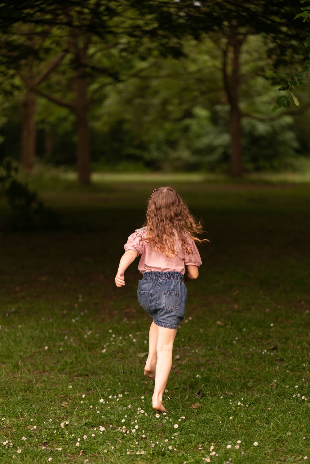 A young girl with curly hair runs barefoot across a green park lawn toward trees during a childhood photography session in Lincolnshire.
