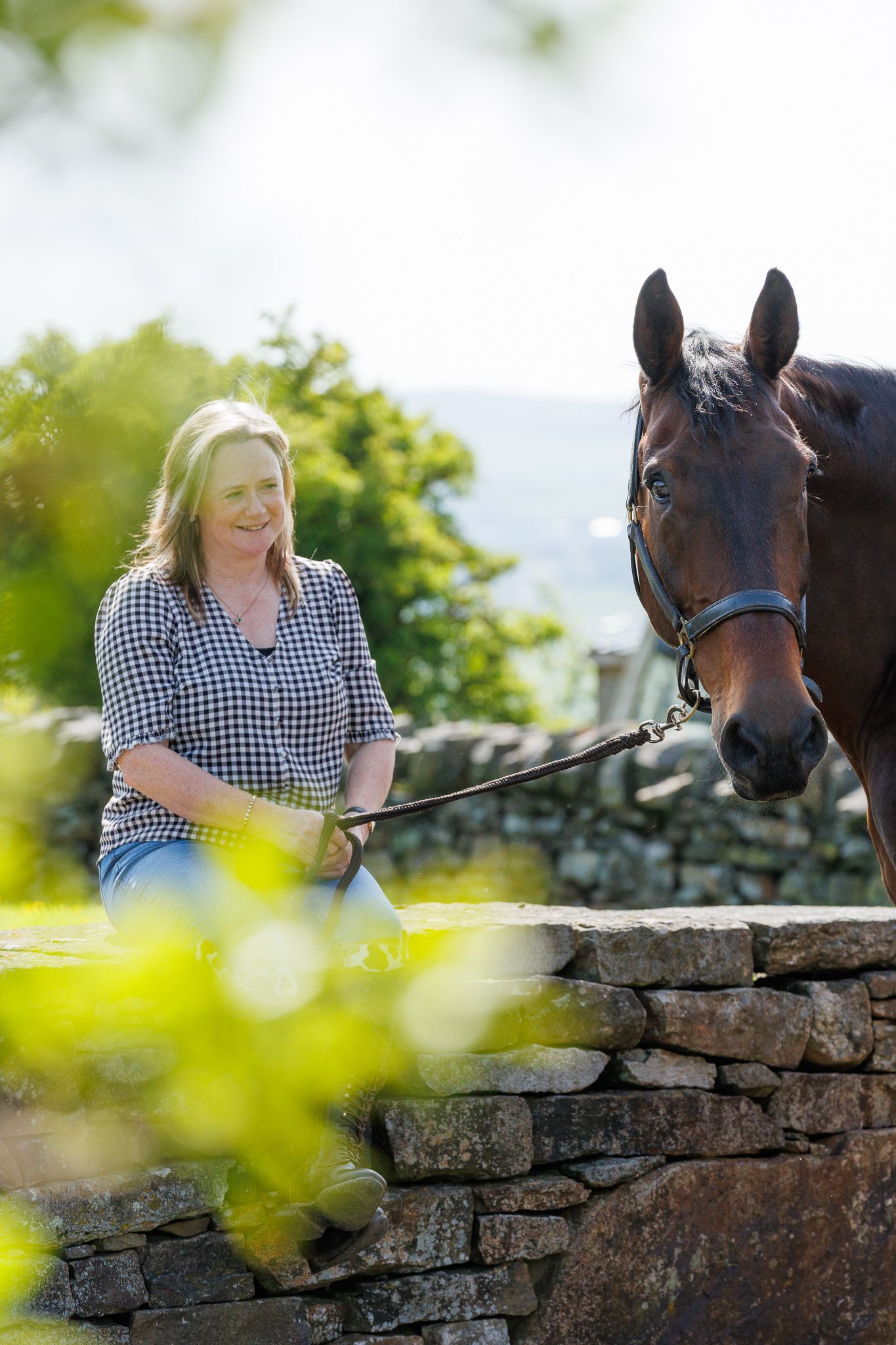 A smiling woman in a plaid shirt holds the reins of a brown horse beside a rustic stone wall outdoors.