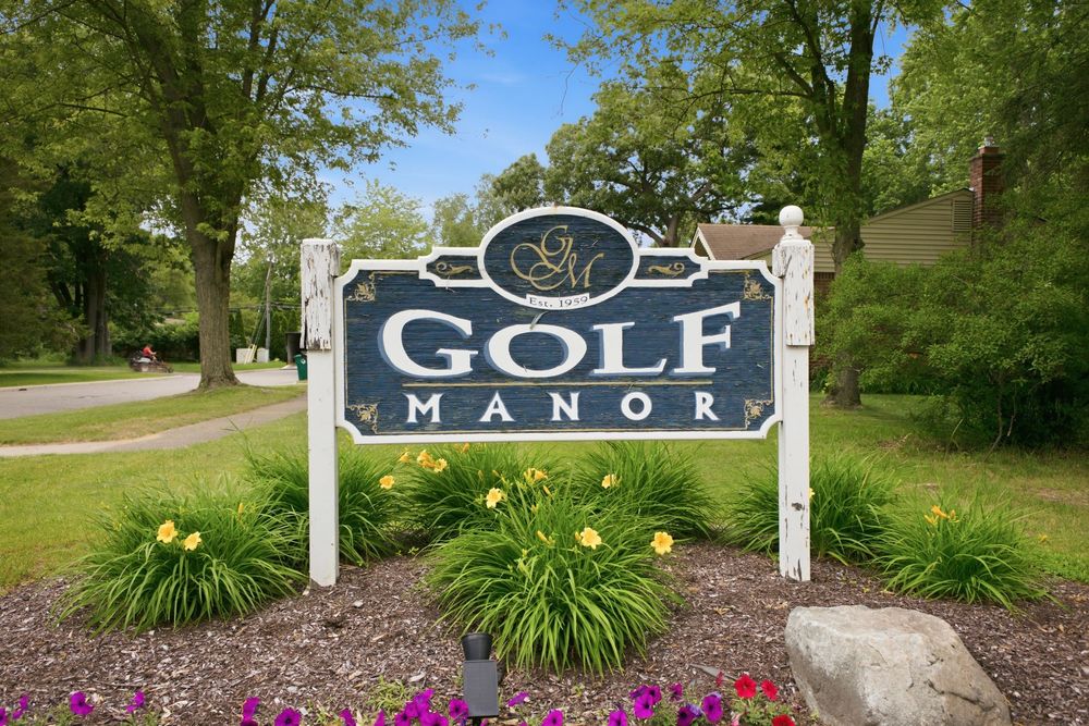Entrance sign for Golf Manor community surrounded by lush green trees, colorful flowers, and manicured landscaping.
