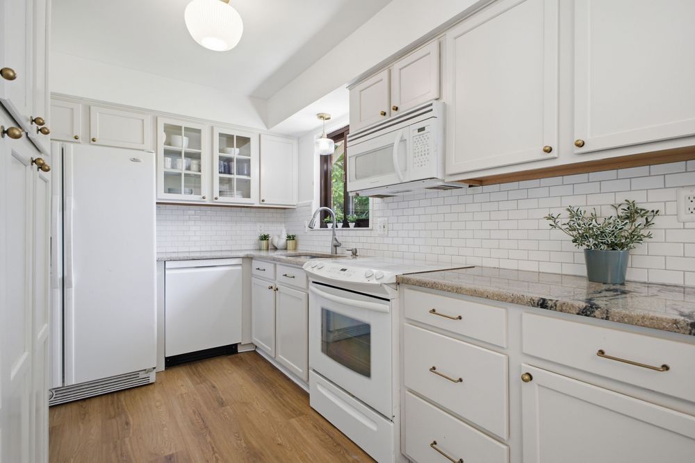 Bright white kitchen with subway tile backsplash, white appliances, glass-front cabinets, and light hardwood floors.