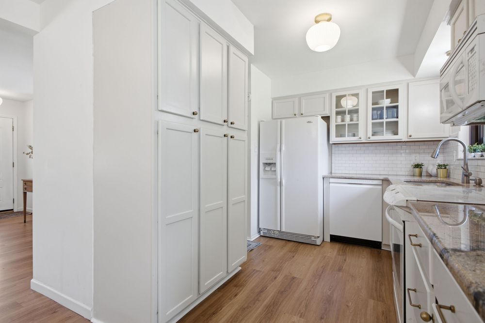 White kitchen with tall pantry cabinets, white refrigerator, glass-front upper cabinets, and granite countertops on hardwood floors.
