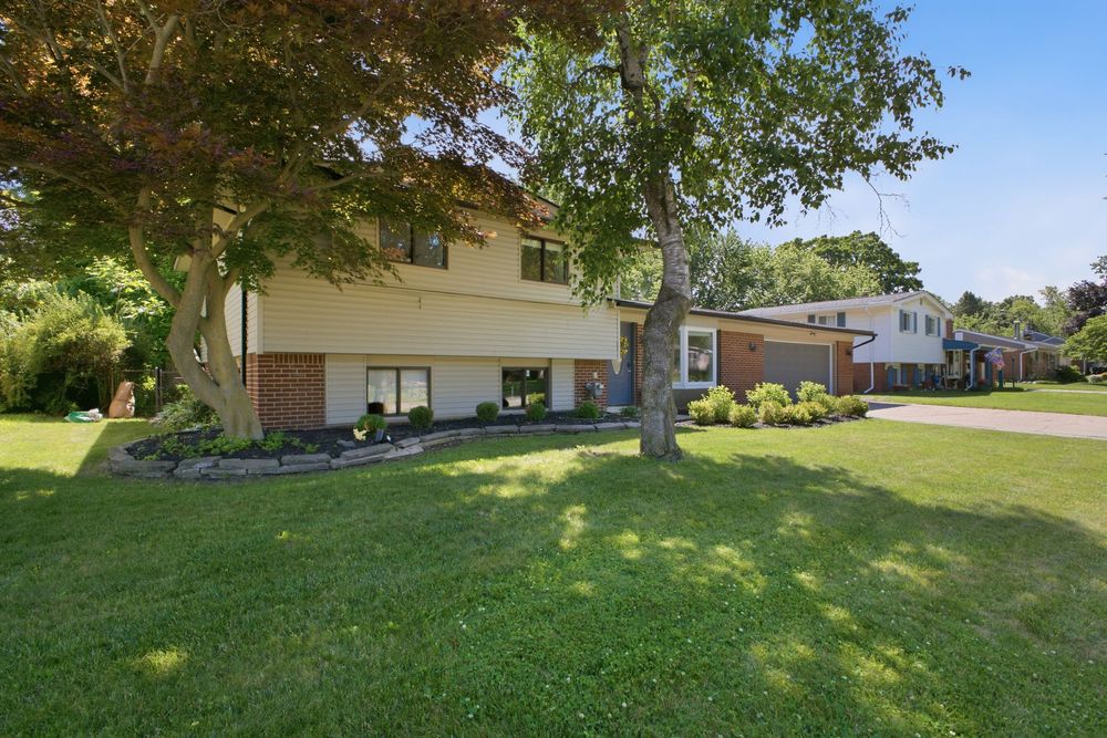 Two-story suburban home with tan siding, large shade trees, and a well-manicured green lawn on a sunny day.