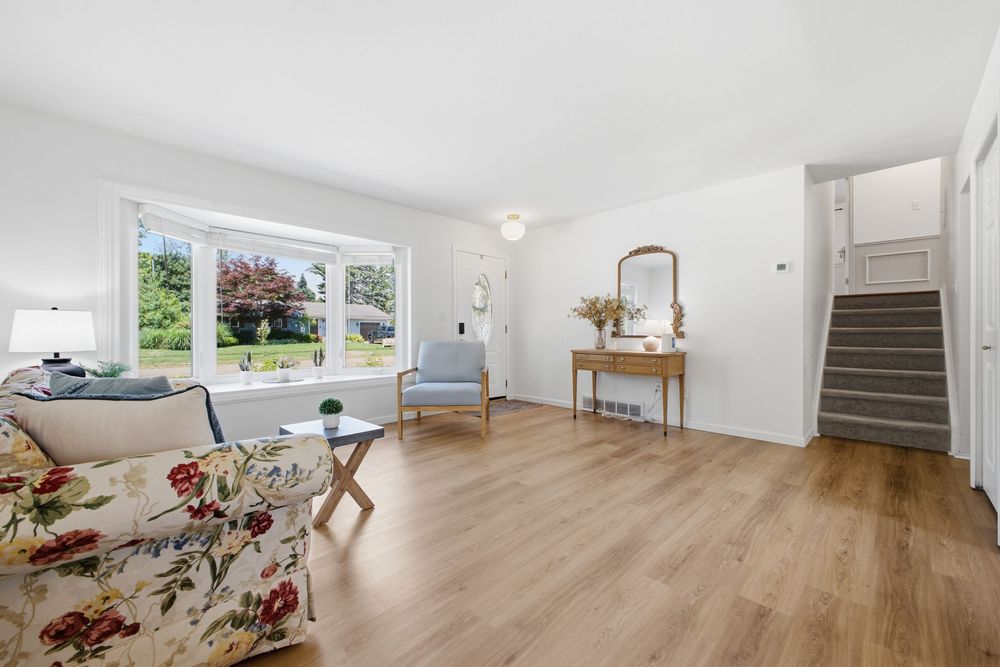 Bright living room with hardwood floors, floral sofa, blue chair, large window, and staircase in a clean white space.