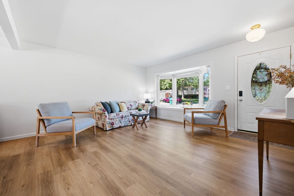 Bright living room with hardwood floors, floral sofa, two blue chairs, and a window letting in natural light.