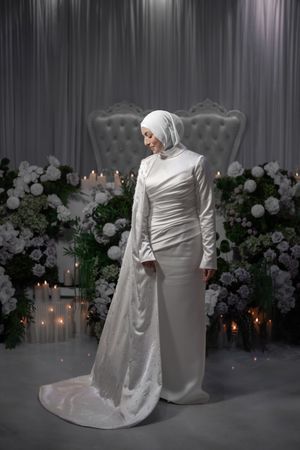 Elegant wedding couple poses in white attire against floral backdrop with ambient lighting.