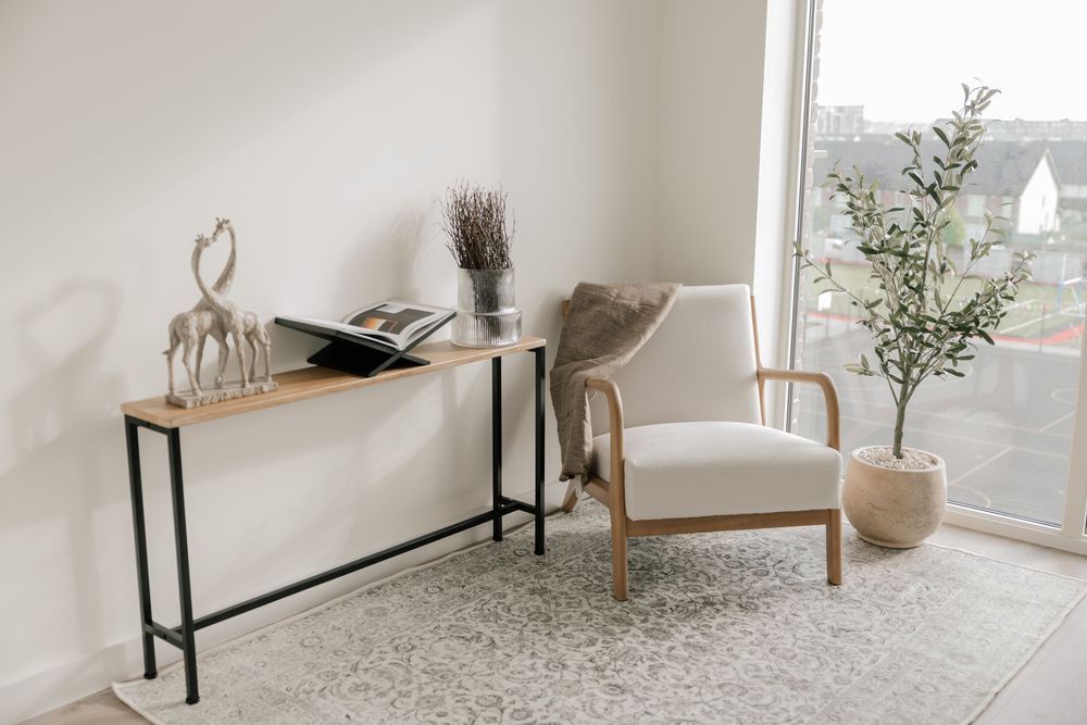 Minimalist living room corner with console table, wooden chair, and potted plant by large window on neutral rug.