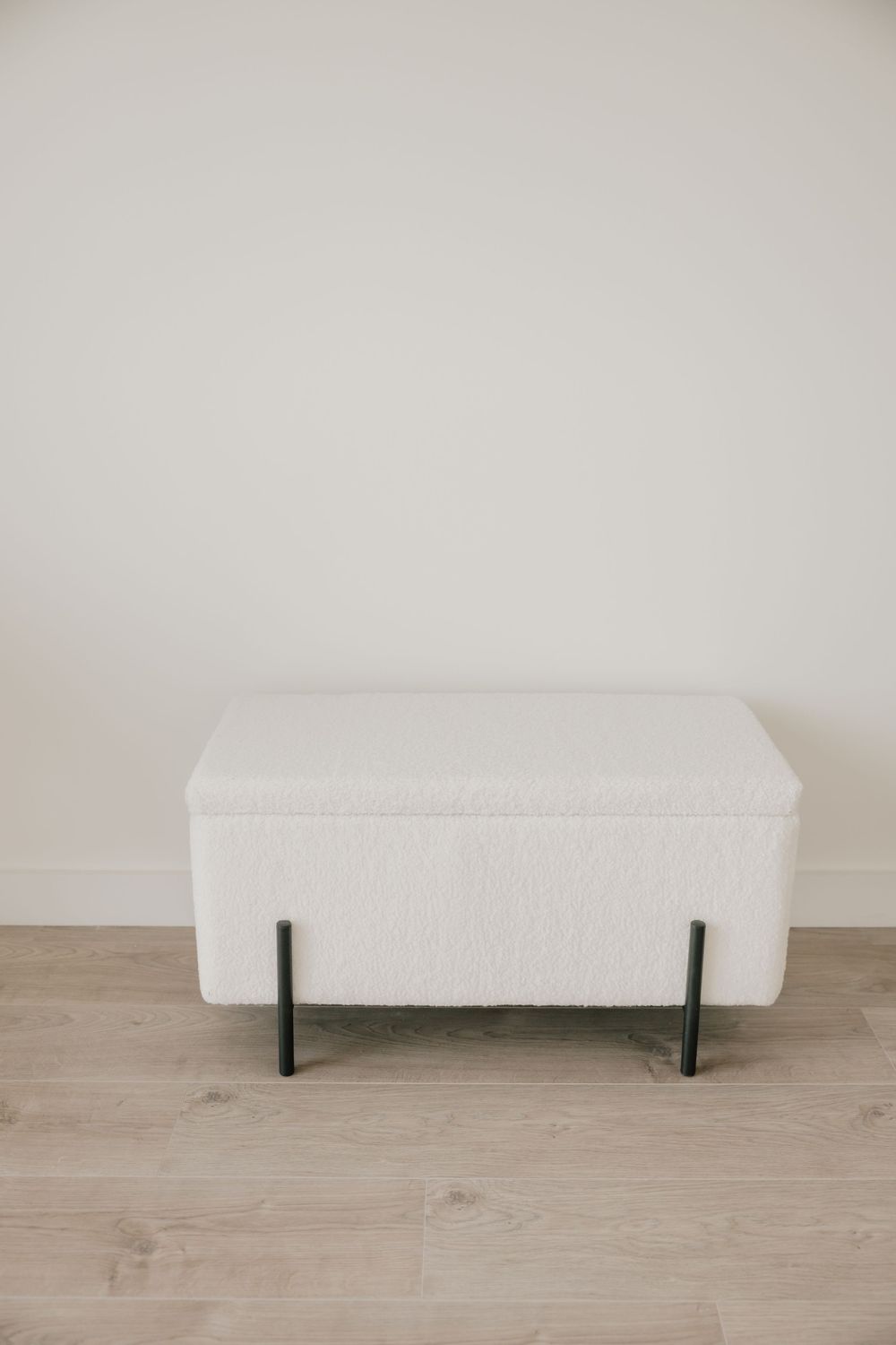 White upholstered storage bench with black metal legs stands against plain wall on light wood flooring.