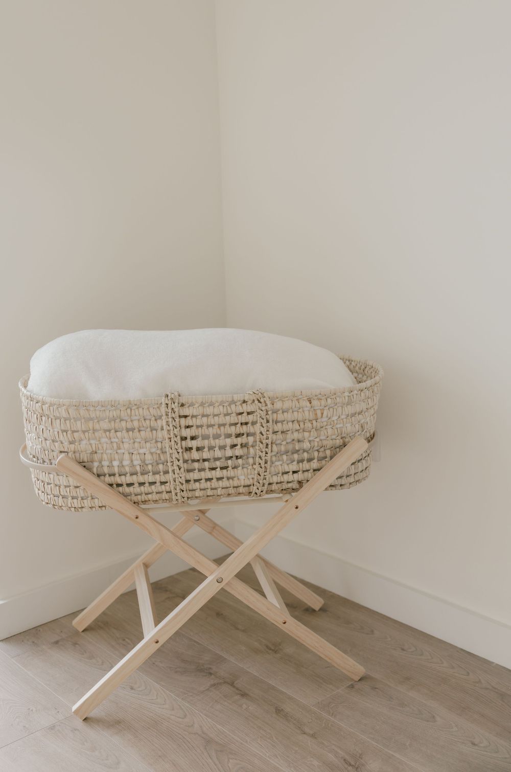 A minimalist woven bassinet with white cushioning sits on wooden cross legs in a corner against light walls.