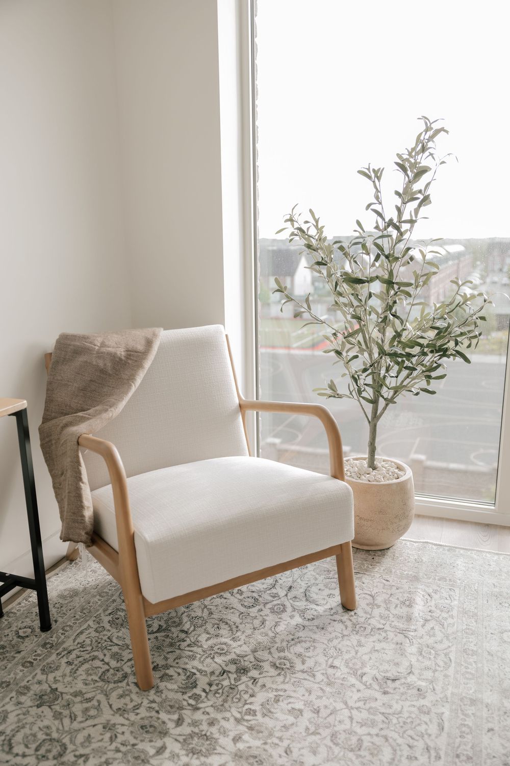 Modern white armchair with wooden frame sits by window with potted olive tree and gray patterned rug in minimalist room.