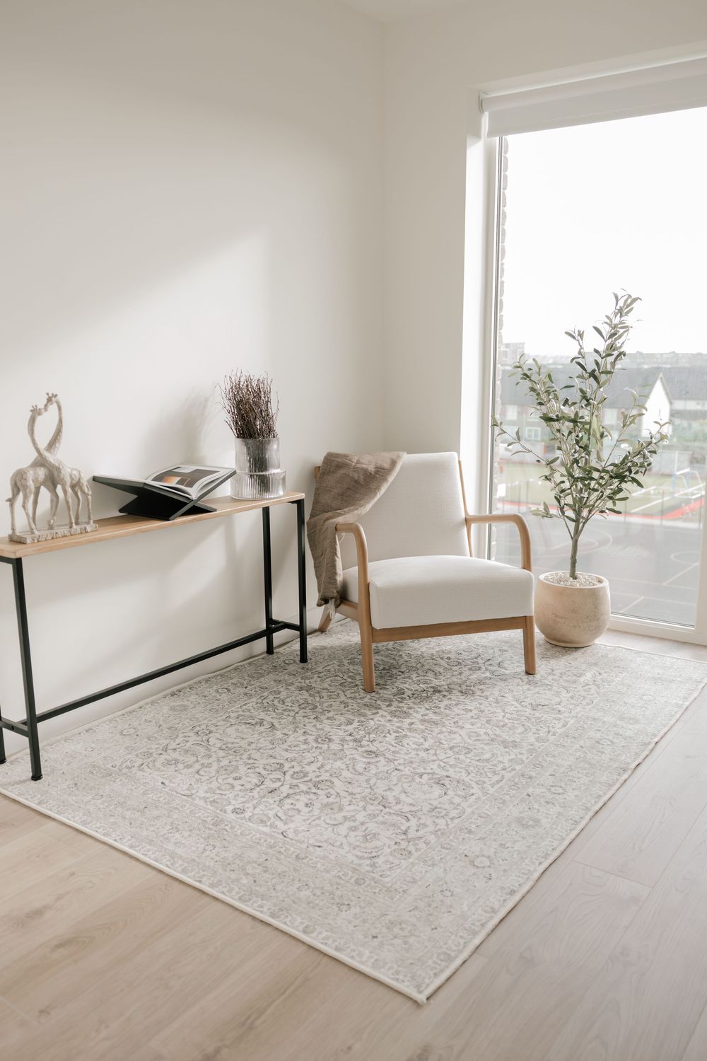 Modern minimalist living room corner with beige armchair, console table, and potted plant by large window and light rug.