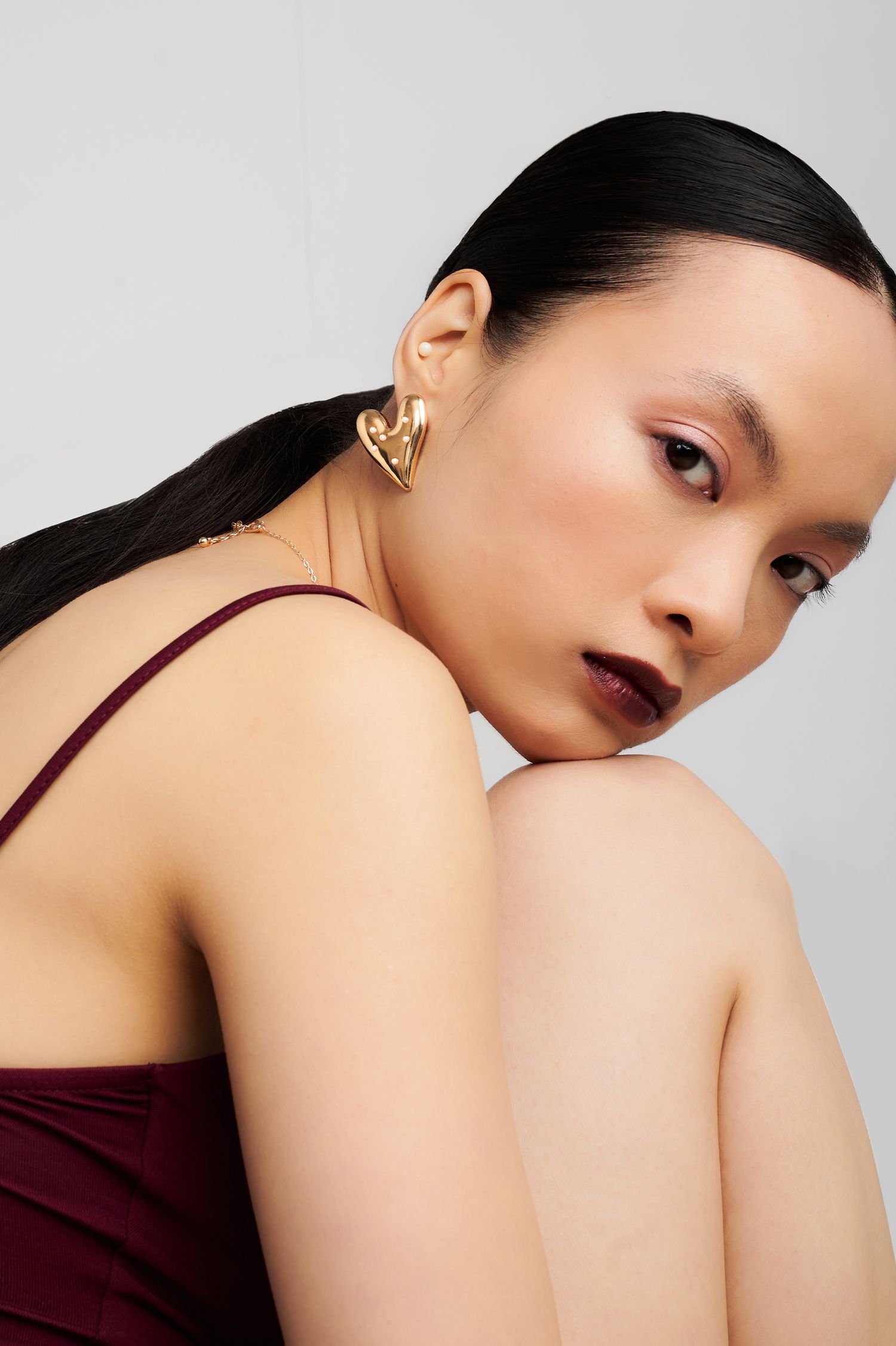 Fashion beauty portrait of woman with dark lipstick wearing burgundy top and gold statement earrings against gray background.