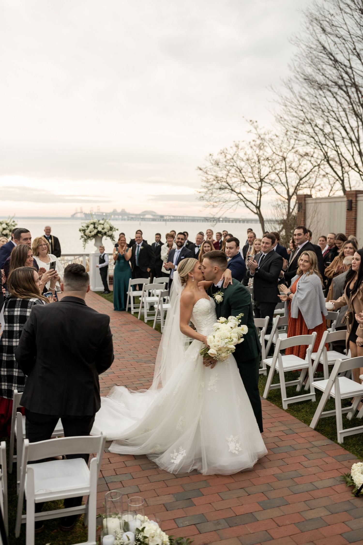 Outdoor wedding ceremony on brick patio with waterfront backdrop and white folding chairs.