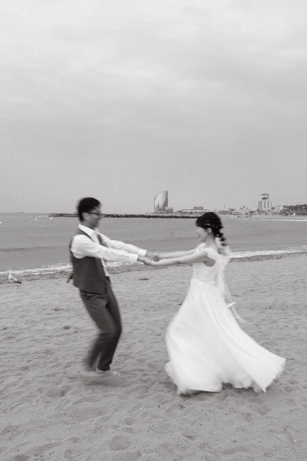 Playful couple in wedding attire dancing and twirling on a sandy beach.