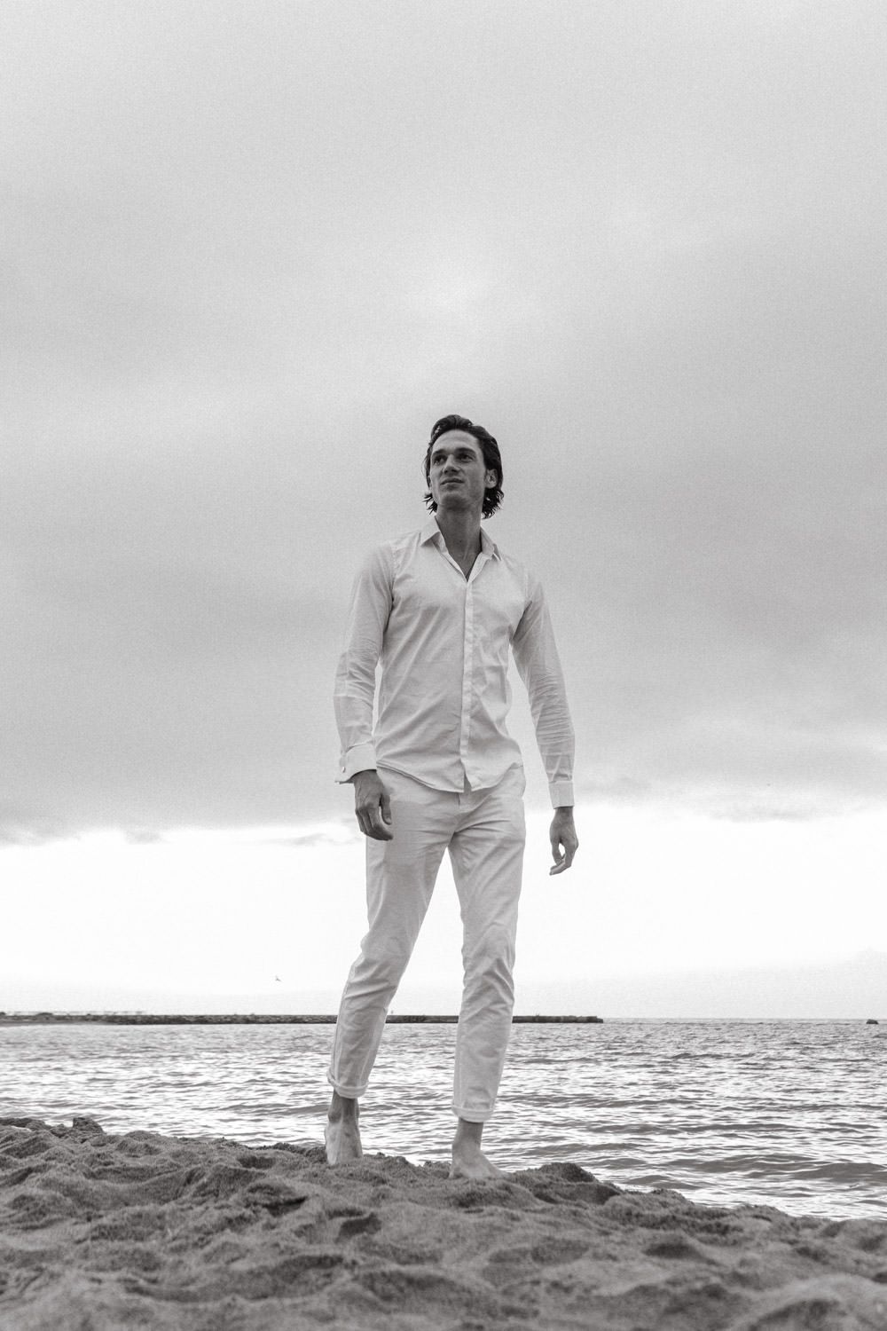 Man in white outfit standing on beach against cloudy sky.