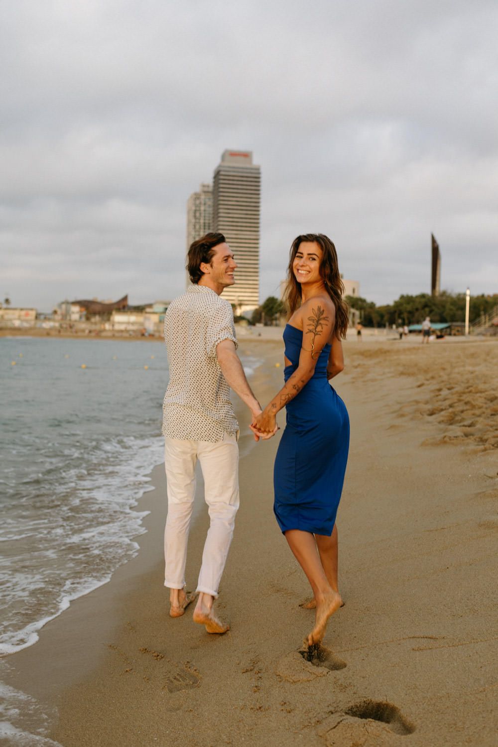 Couple holding hands while walking on beach with city skyline in background.