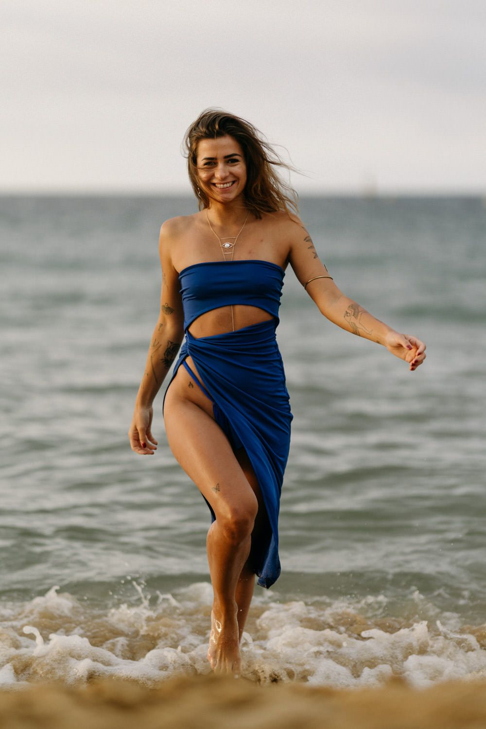 Woman in blue beach dress poses playfully in ocean waves at sunset.