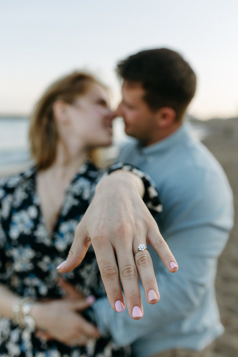 Close up of engagement ring on beach with couple embracing in background.