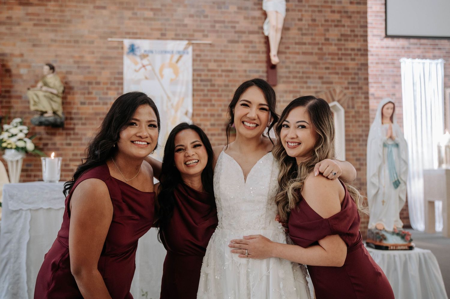 Four people wearing burgundy and white dresses pose together at a wedding reception with brick walls and decorations.