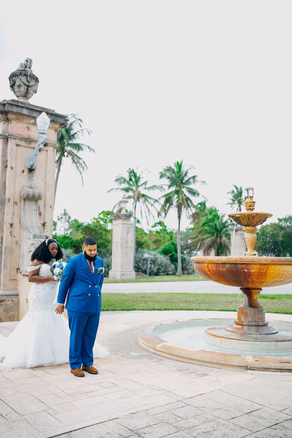 A couple in wedding attire poses near an ornate stone fountain and palm trees at a tropical venue.