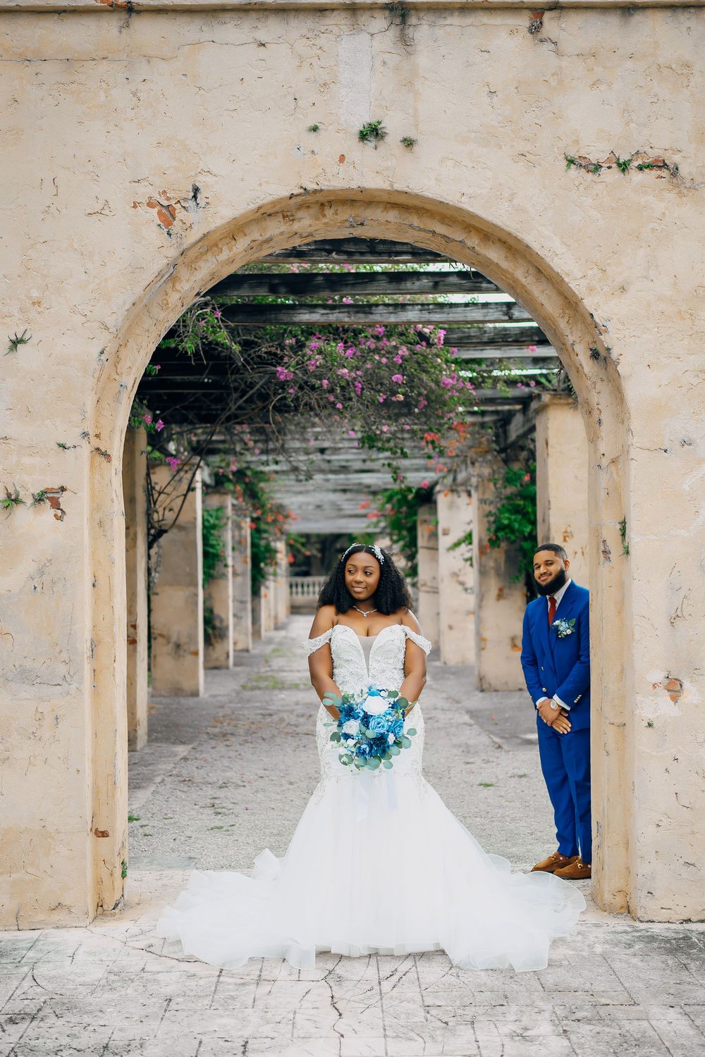 Newlyweds stand under an arched passageway with climbing flowers, bride holding blue bouquet in mermaid gown.