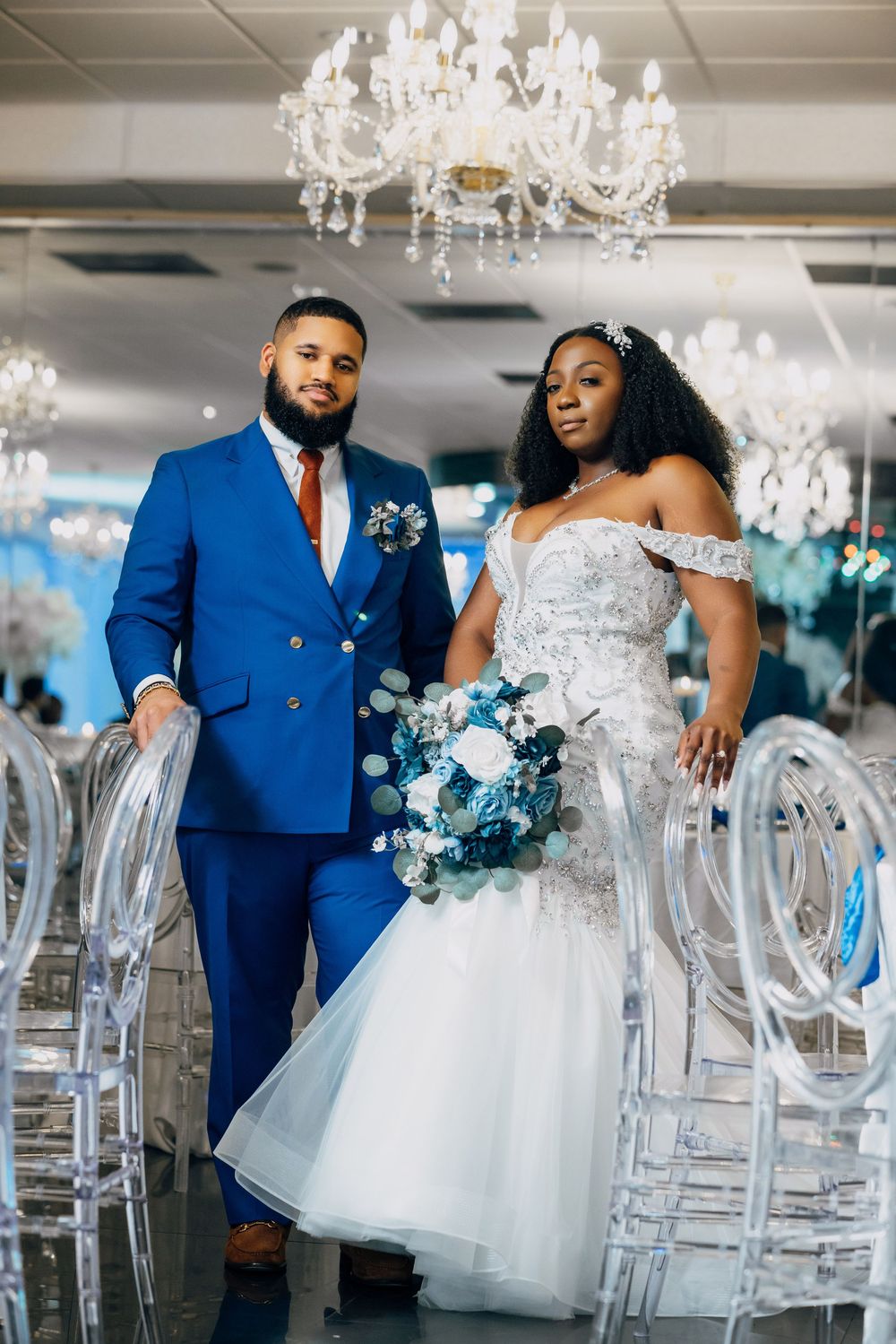 A couple in blue and white wedding attire stands beside crystal chairs under an elegant chandelier.