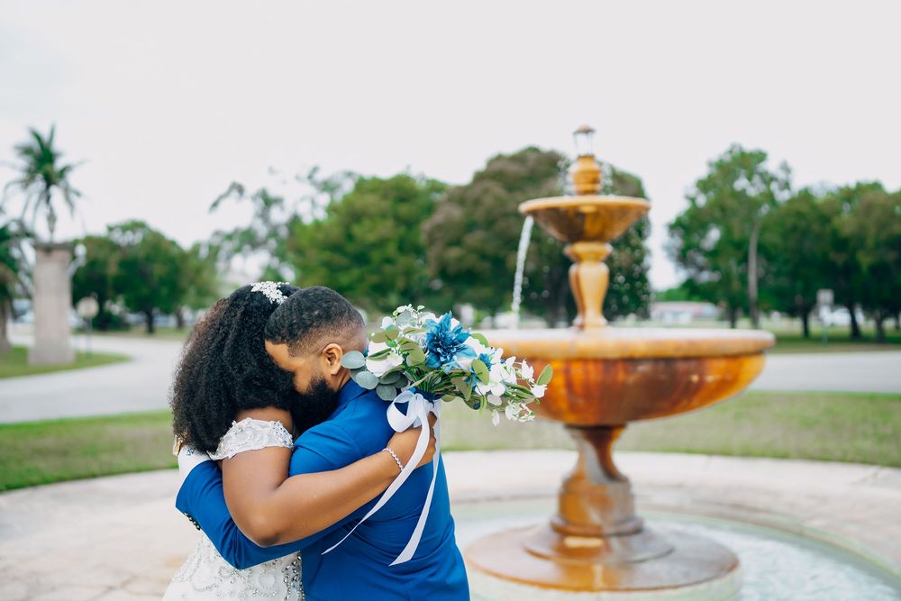 Two people embrace near an ornate copper fountain in a park setting with palm trees.