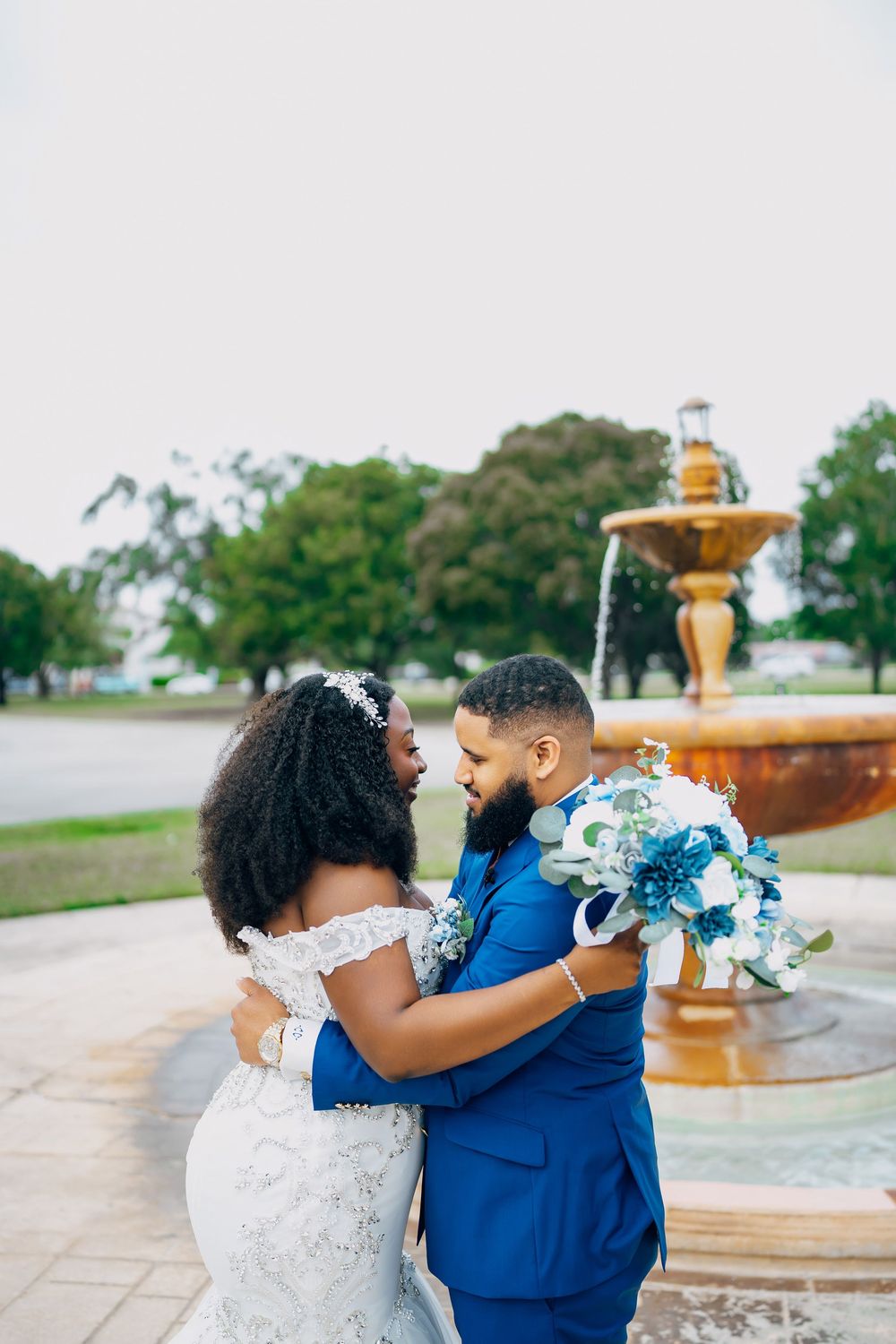 A couple embraces near an ornate fountain during their wedding, with the bride holding a blue and white bouquet.