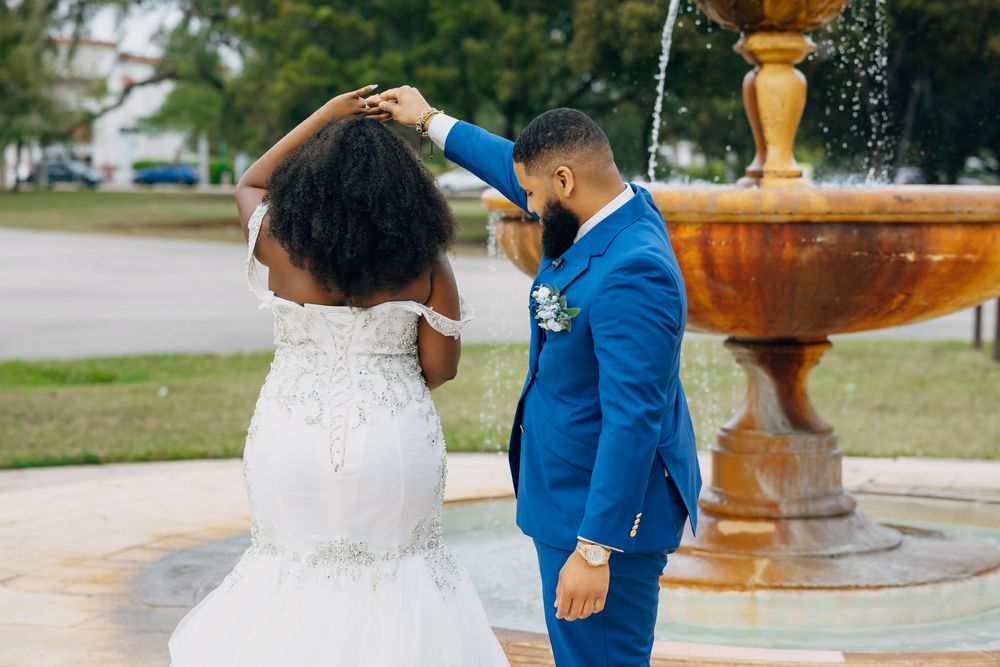 Bride in lace gown and groom in blue suit share a romantic moment by a decorative fountain.