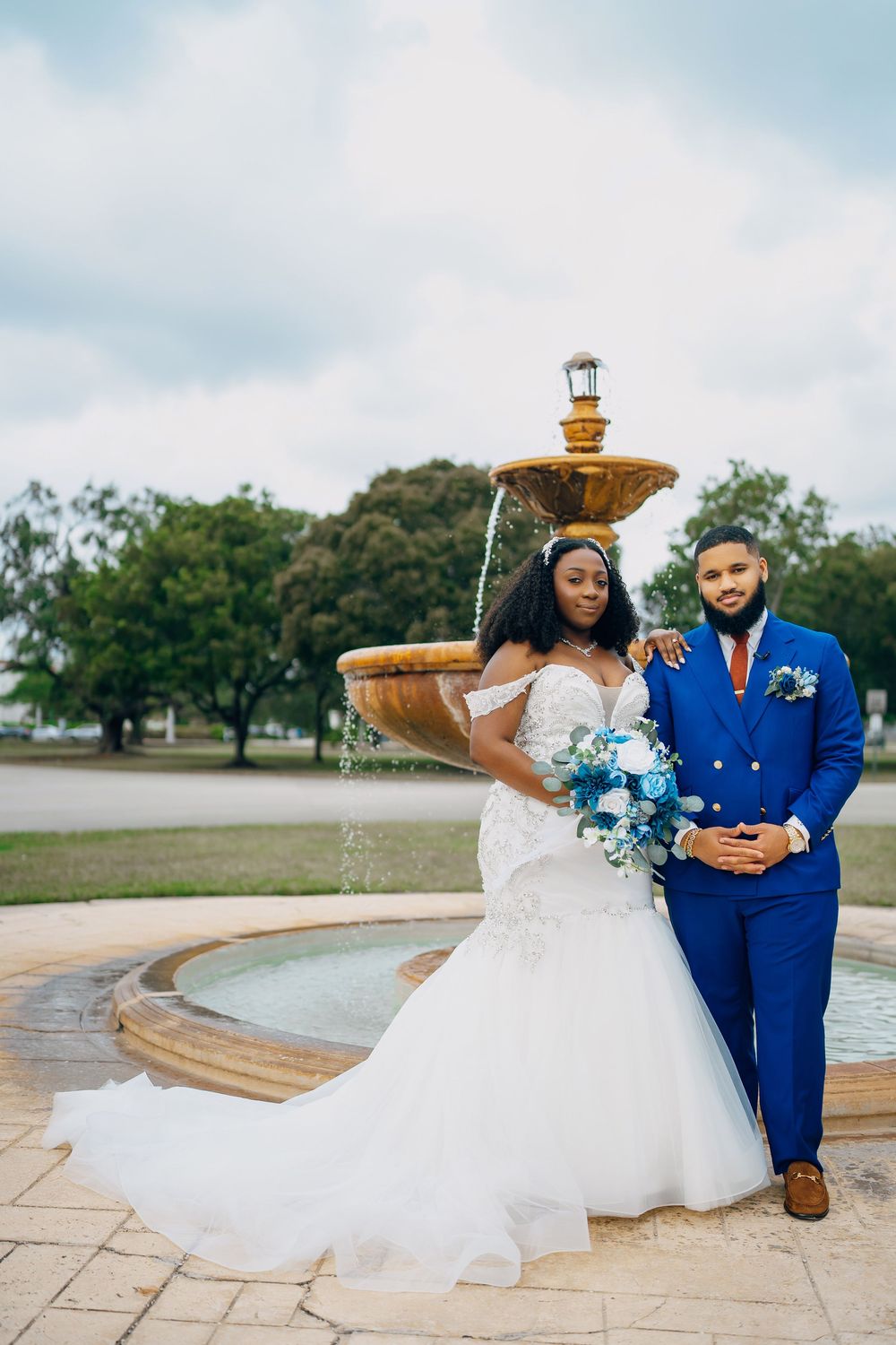 Newlyweds in royal blue suit and white gown pose elegantly by a tiered fountain in a garden setting.