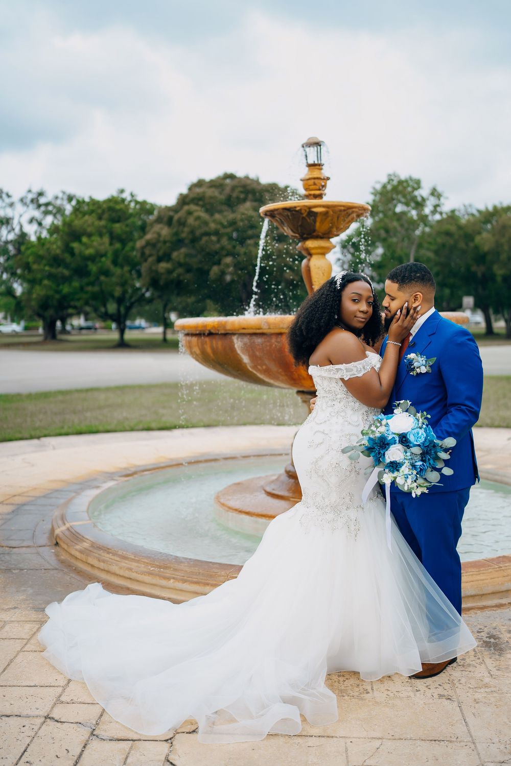 A wedding couple in elegant attire embrace by an ornate stone fountain in a garden setting.
