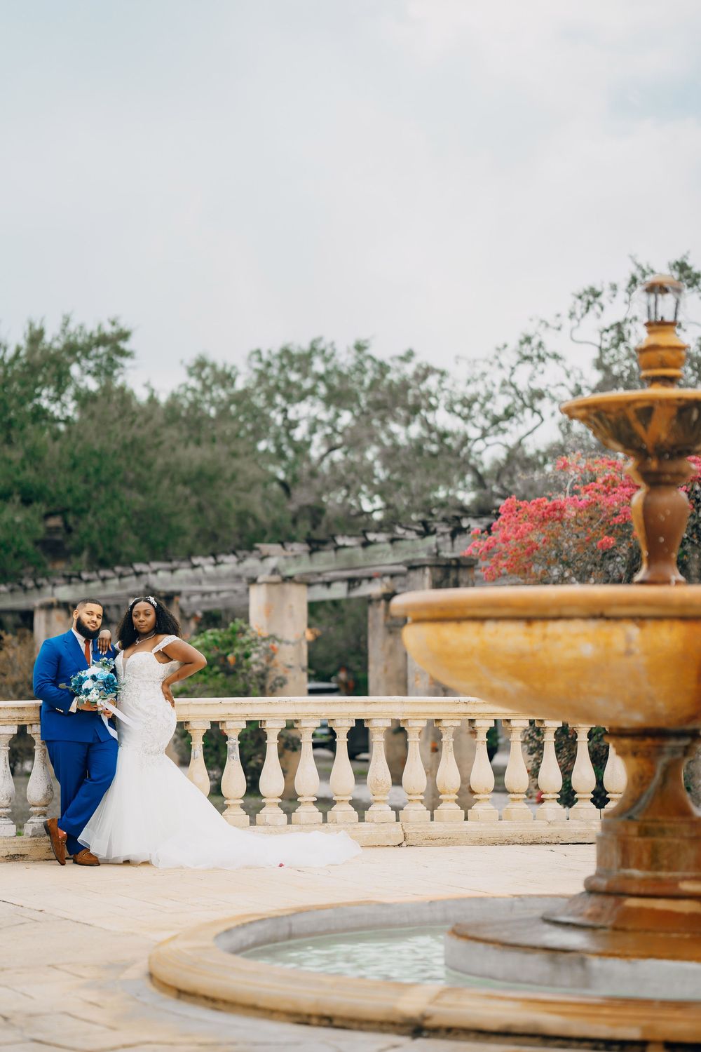 A couple in wedding attire pose near a vintage fountain with pink flowers and stone balustrades in the background.