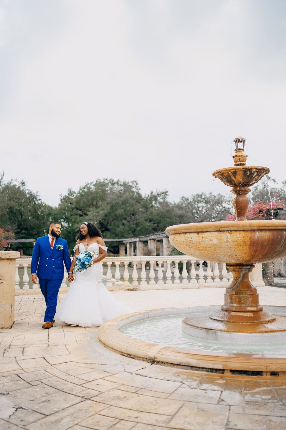 Elegant couple walking near a grand stone fountain in a traditional courtyard setting.