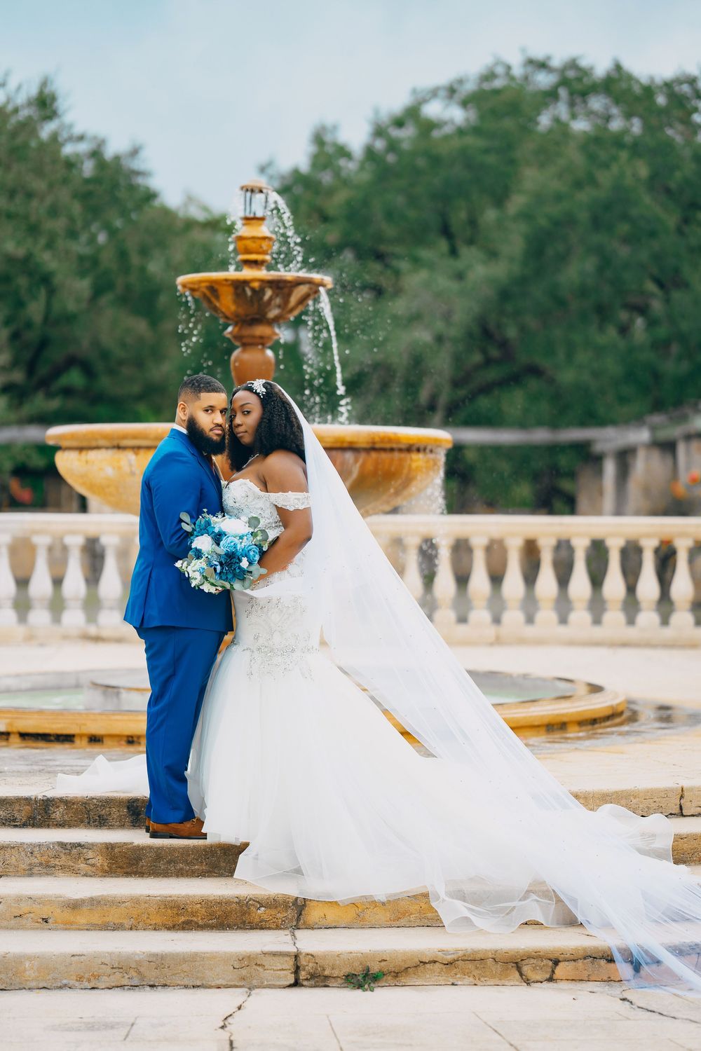 Wedding couple stands on stone steps near a flowing fountain, with bride's dramatic veil trailing behind.