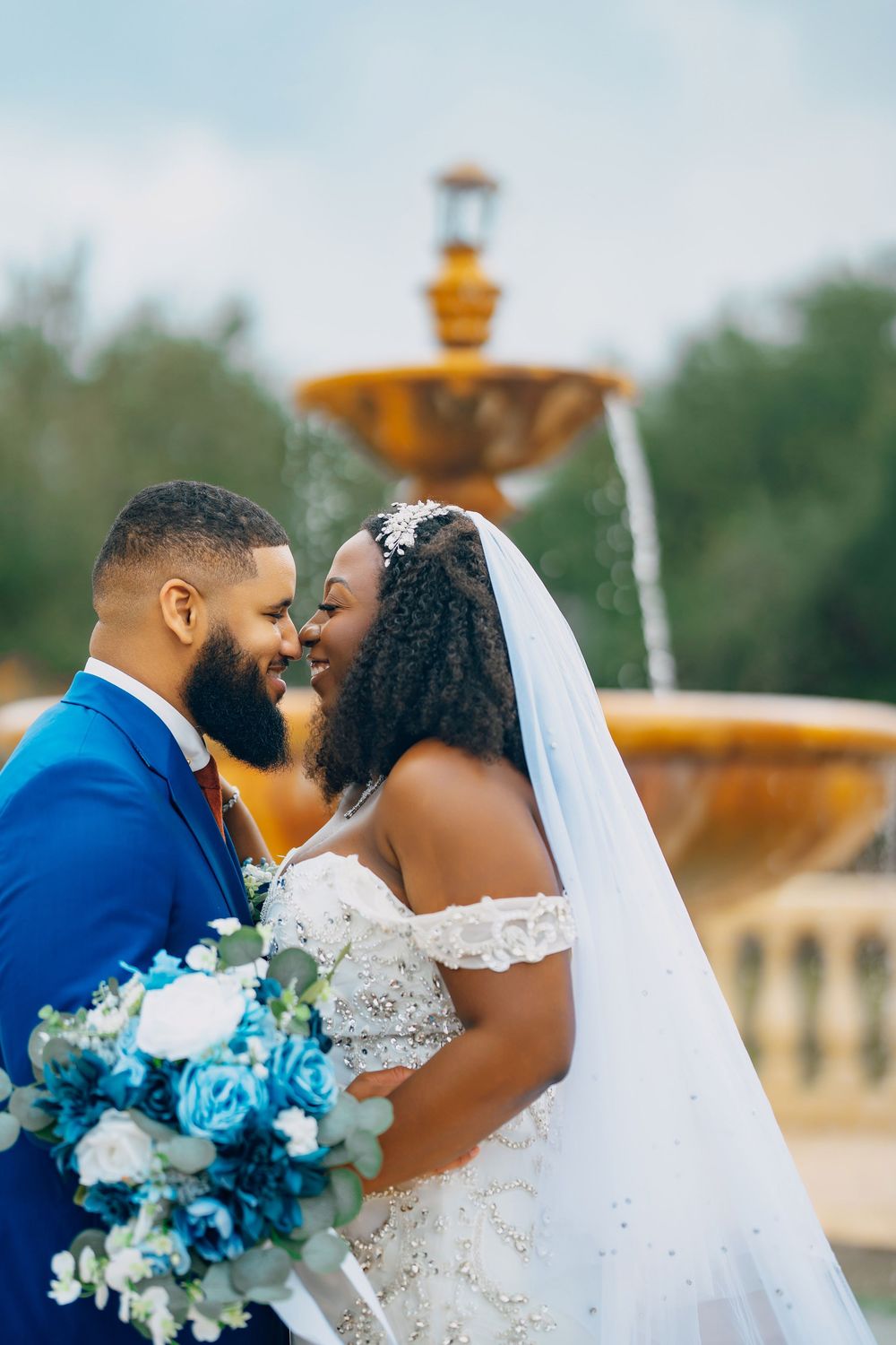 A couple embraces during their wedding photo session by a golden fountain with blue and white bouquet.