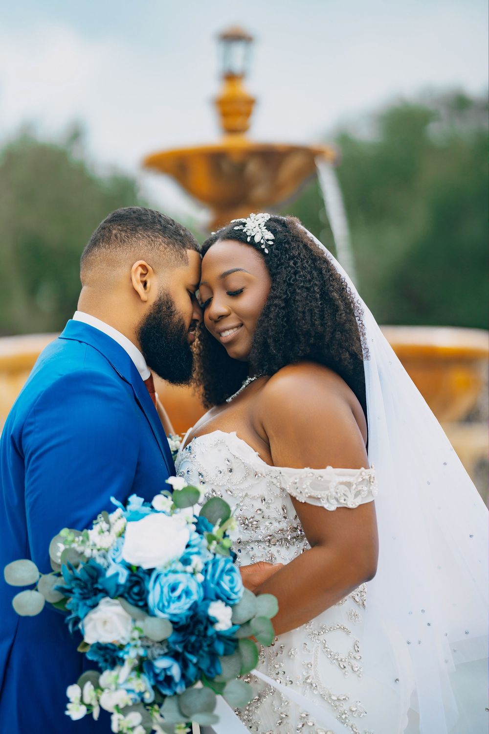 Intimate wedding portrait of couple sharing tender moment by fountain with blue and white bouquet.