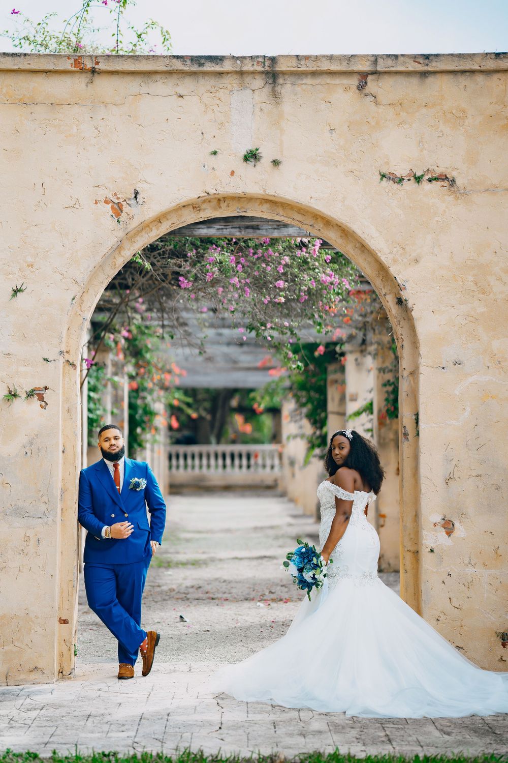 A couple poses in a romantic stone archway with flowering vines, the bride in white and groom in royal blue suit.