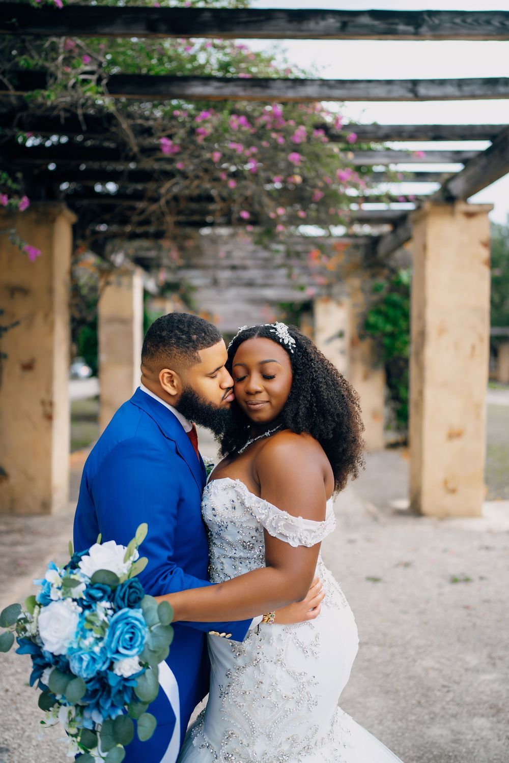 Romantic couple embraces under a pergola with climbing flowers during their tropical garden wedding.
