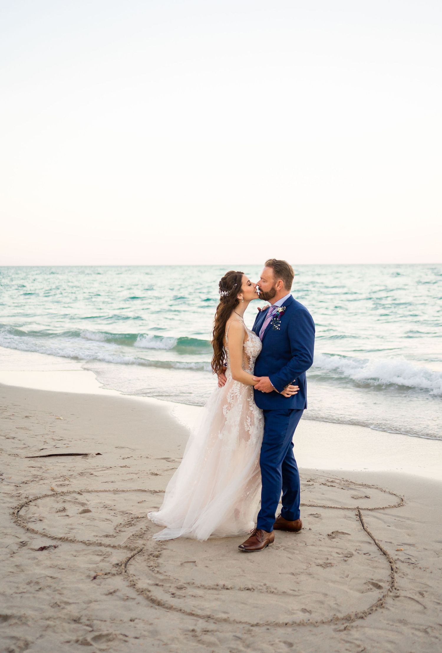 Couple shares romantic moment on beach at sunset with heart drawn in sand and waves in background.