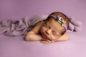Sleeping newborn baby wearing delicate floral headband rests peacefully on soft purple backdrop during professional photoshoot.