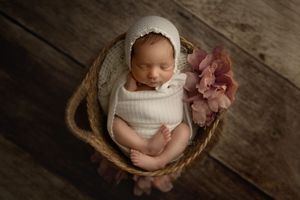 Newborn baby resting in wooden basket with delicate pink flower decoration.