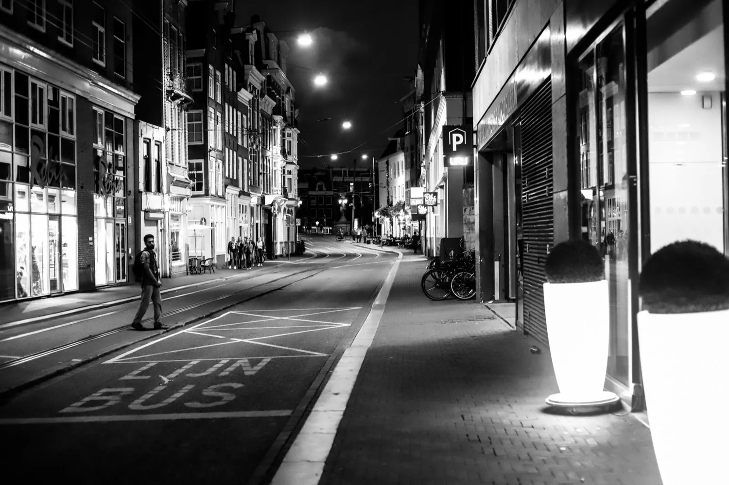 Scène urbaine de nuit en noir et blanc, avec un piéton marchant dans une rue éclairée par des lampadaires.