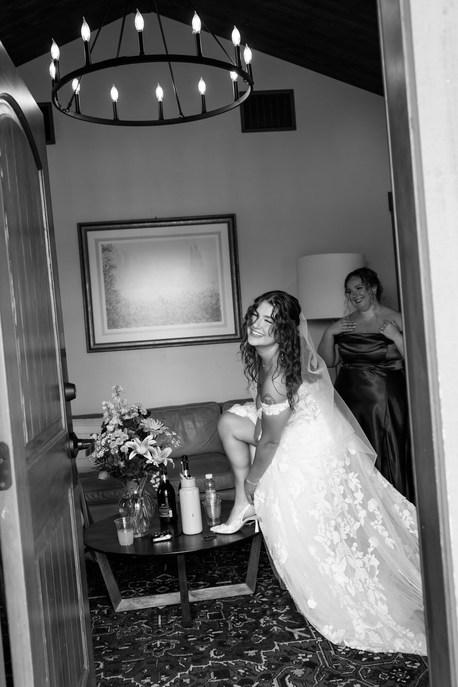 Black and white wedding photograph showing a figure in a lace wedding dress and someone in a dark gown under a lit chandelier.
